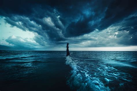 Girl Standing At Edge Of Sea Under Dark Clouds By Tj Drysdale
