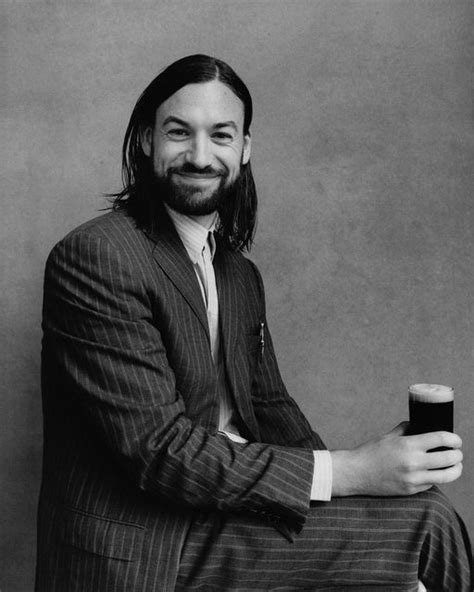 a man with long hair sitting down holding a beer in his hand and smiling at the camera