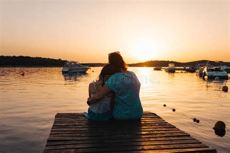 Mom And Daughter Sitting On A Wooden Pier Hugging Each Other Admiring The Setting Sun Over The