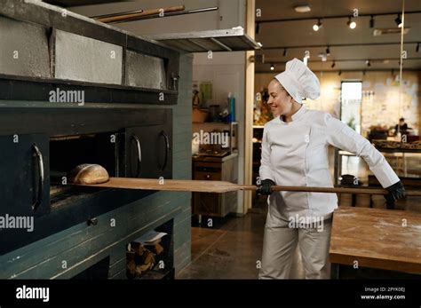 Young Bakery Worker Using Wooden Shovel To Take Bread From Stove Oven