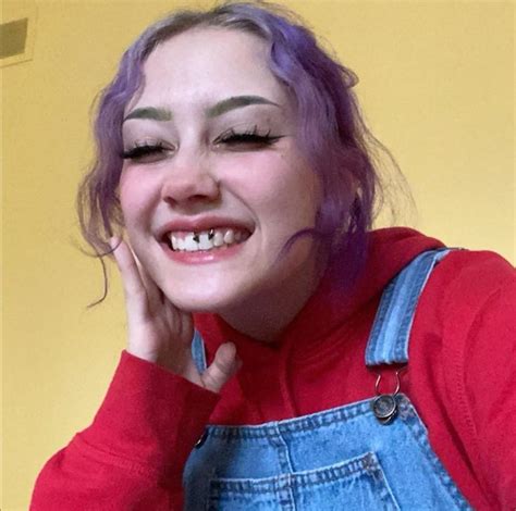 A Woman With Purple Hair And Blue Overalls Smiles While Holding Her Hand To Her Face