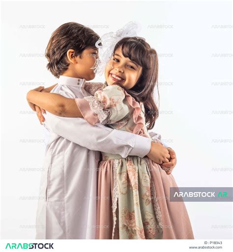 خلفية بيضاء لشقيق وأخته صغيران سعوديان مبتسمان ، يعانقان بعضهما وينظر الأخ لإخته الصغيرة