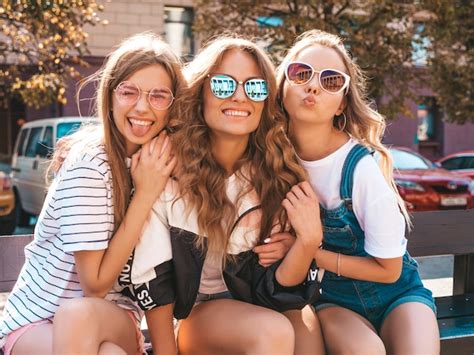 トレンディな夏服の3人の若い美しい笑顔流行に敏感な女の子の肖像画。通りのベンチに座っているセクシーな屈託のない女性。サングラスで楽しんでいる
