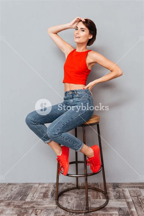 full length portrait of a smiling cute woman sitting and posing on the chair over gray