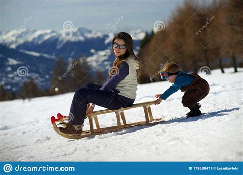 Young Smiling Woman On Sled On Snowy Slope And Little Child Pushing
