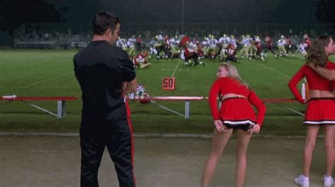 Two Cheerleaders Are Standing In Front Of A Football Field While An Official Looks On