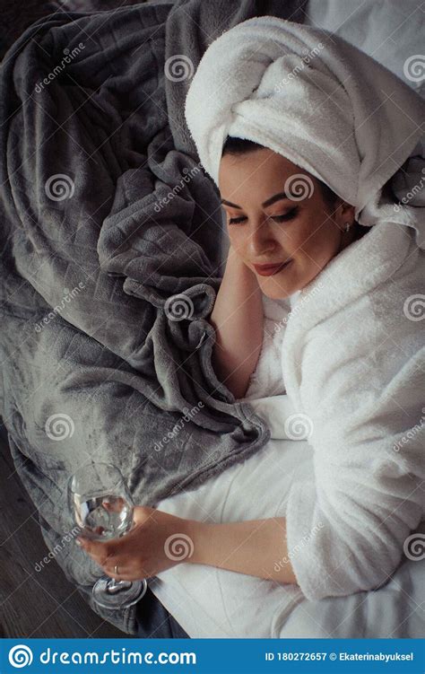 Young Woman In A Bathrobe And A Towel On Her Head Lies On The Bed In A Glass Of Water In Her