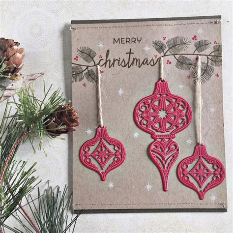 A Christmas Card With Two Ornaments Hanging From Its Side And Pine