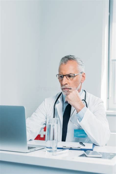 Serious Middle Aged Male Doctor Using Laptop At Table Stock Photo