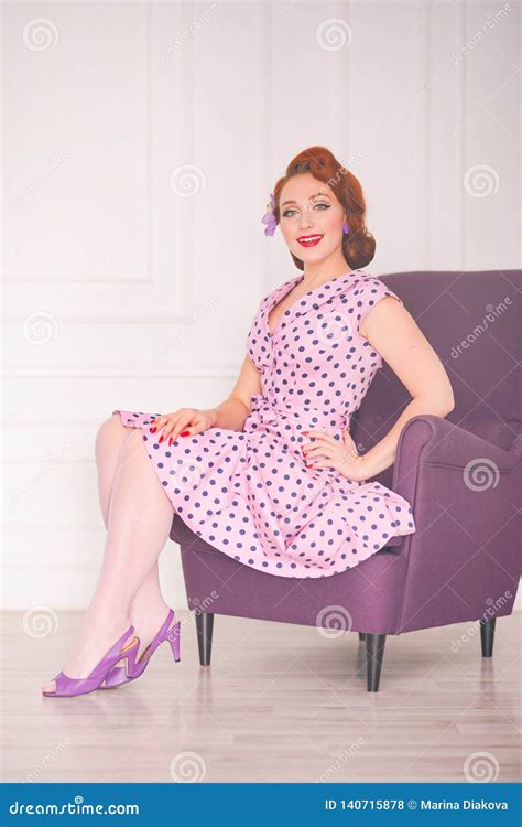 Pretty Redheaded Pin Up Woman Wearing Pink Polka Dot Dress And Posing With Purple Armchair On
