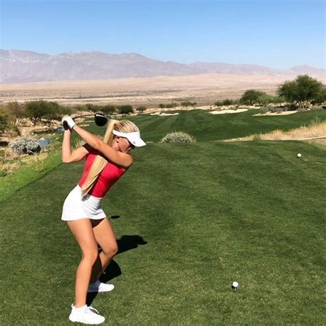 A Woman Swinging A Golf Club On A Green Field With Mountains In The