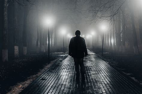 Sad Man Alone Walking Along The Alley In Night Foggy Park Back View