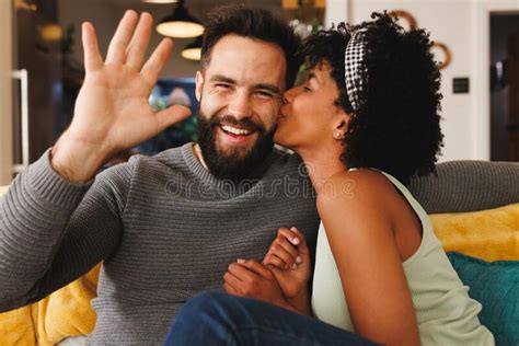Happy Bearded Biracial Man Waving Hand And Girlfriend Kissing Him On Cheek While Relaxing On