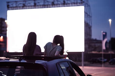 büyük beyaz ekranlı bir açık hava sinemasında film izlerken arabada oturan iki kadın arkadaşın