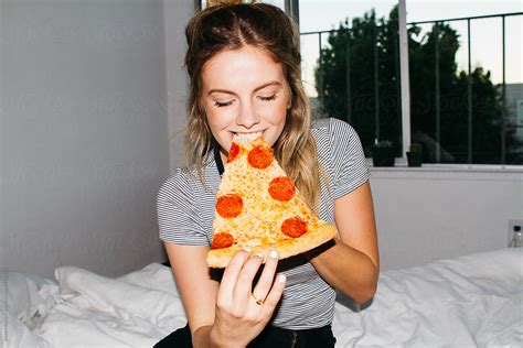Young Woman Eating Slice Of Pepperoni Pizza On Bed In Apartment By