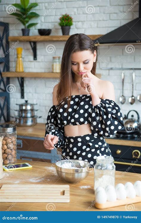 Beautiful Young Brunette Girl In A Polka Dot Dress Looks At The Kitchen Recipe Cake Dishes Stock
