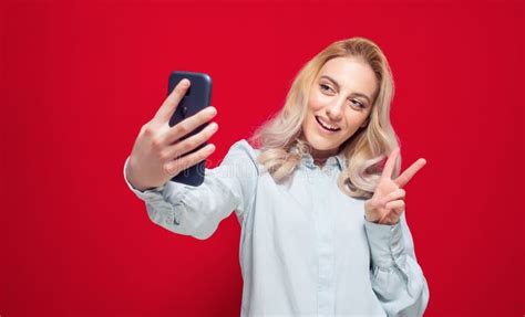 Trendy Girl Holding In Hands Smartphone And Making Selfie While Showing V Sign Isolated On Red