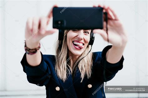 Playful Woman Sticking Out Tongue While Taking Selfie Against Wall — One Person Headphones