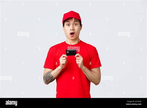 Nervous Asian Delivery Guy In Red T Shirt And Cap Uniform Holding Credit Card Staring Camera