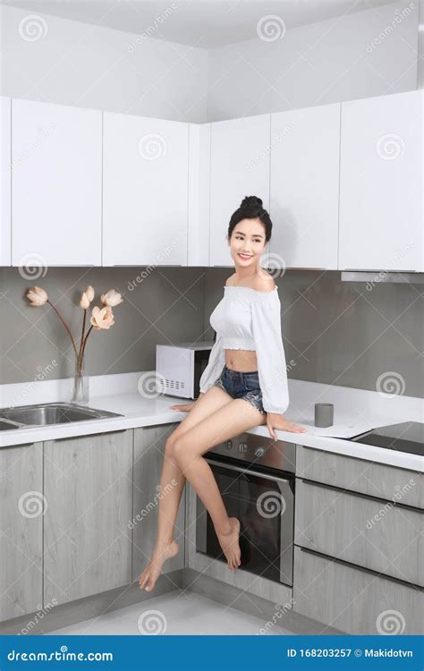 Cheerful Woman Sits On Countertop In White Modern Kitchen Morning Time