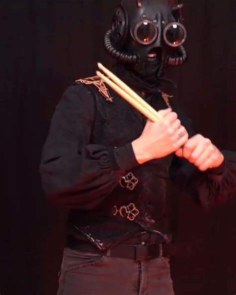 A Man In A Gas Mask Holding Two Drumsticks And Wearing A Black Outfit