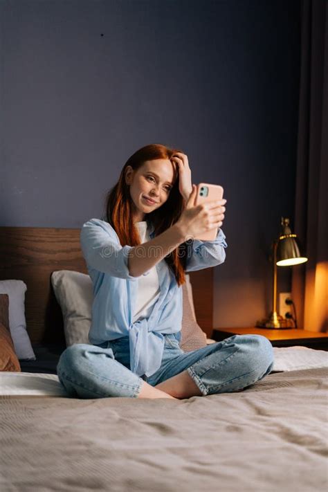 Attractive Redhead Young Woman Posing For Mobile Selfie In Bed While Sitting On Bed At Cozy