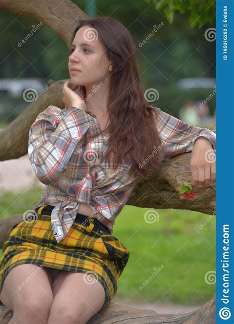 Slender Brunette Girl In A Plaid Skirt And Blouse In The Summer By A