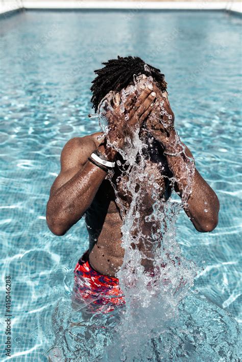 Anonymous African American Male In Shorts Standing In Swimming Pool And Splashing Water In Face