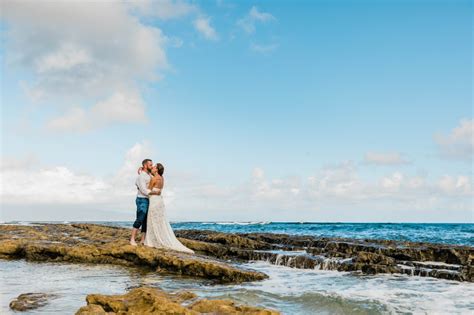 A Bride And Groom Standing On The Rocks By The Ocean With Their Arms Around Each Other