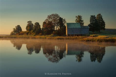 A Small House Sitting On The Shore Of A Lake At Sunset With Trees In