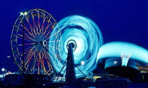 The Ferris Wheel Is Lit Up At Night In Front Of Some Other Amusement