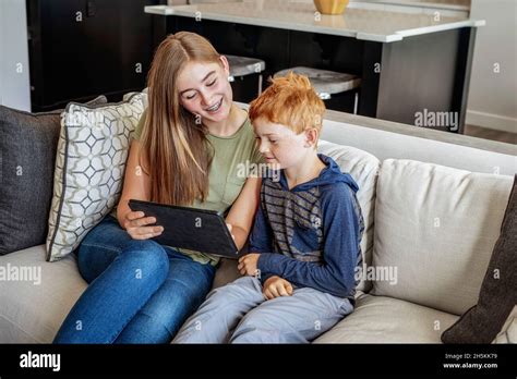 A Brother And Sister Sit On A Couch At Home With A Tablet And Viewing