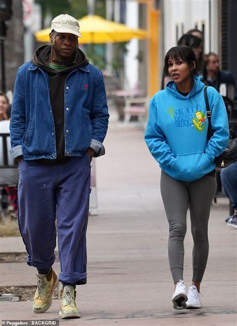 A Man And Woman Walking Down The Street With People Sitting On Benches