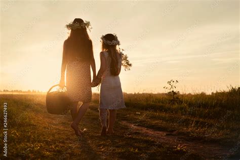 Two Girls Hold Hands And Walk Across The Field Barefoot At Sunset