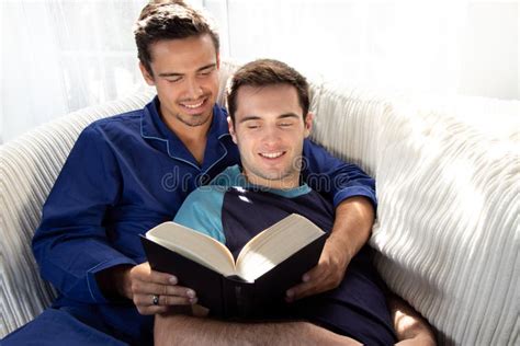 male gay couple cuddling and reading together on large couch next to patio doors with bright