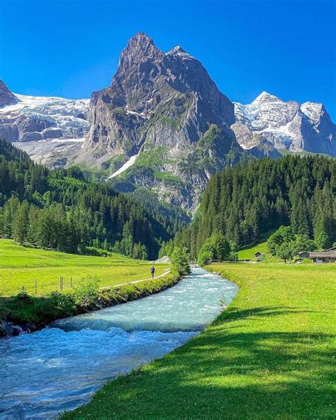 A River Running Through A Lush Green Field Next To Tall Snow Covered