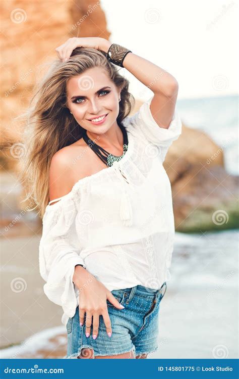 Pretty Blonde Girl With Long Hair Is Posing To The Camera On The Beach Near Sea She Wears White