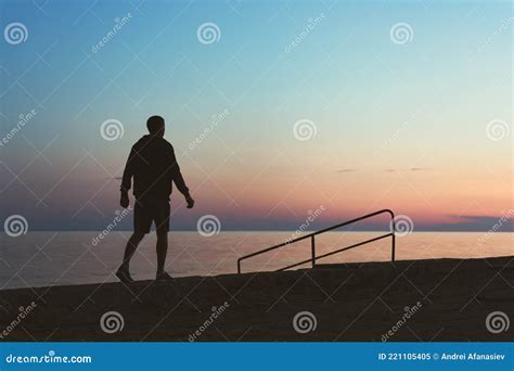 Silhouette Of A Young Man Walking Along The Embankment At Sunset Stock