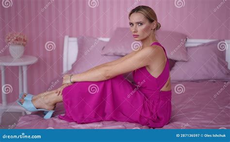 Side View Portrait Of Slim Young Woman In Pink Dress On Pink Bed With Serious Facial Expression