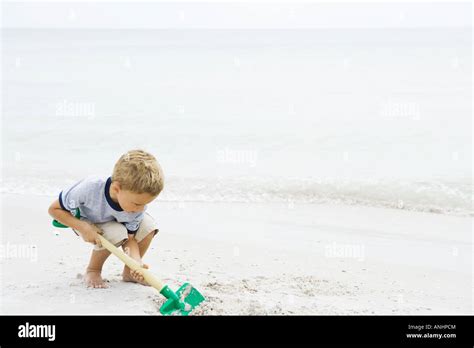 Young Boy Crouching At The Beach Digging In Sand With Shovel Stock