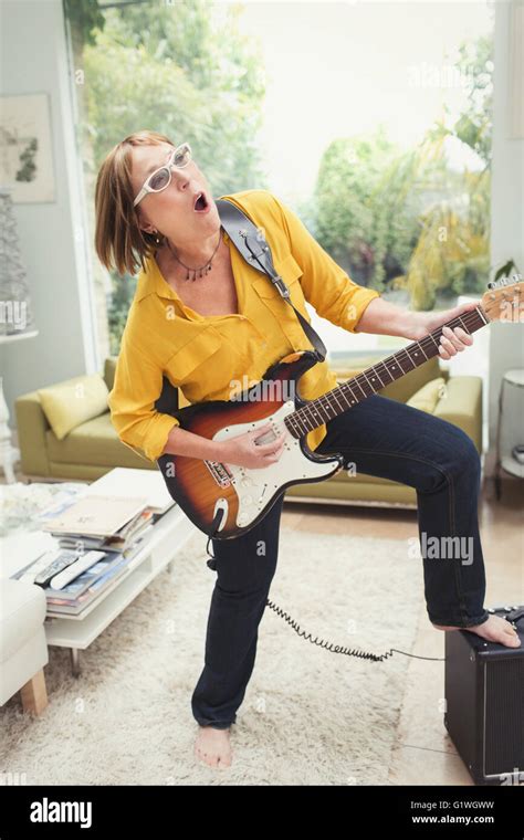 Playful Mature Woman Playing Electric Guitar In Living Room Stock Photo