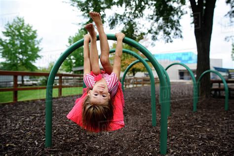 Portrait Of Girl Hanging Upside Down On Outdoor Play Equipment At Playground Stock Photo