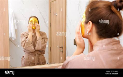 brunette woman in brown bathrobe with golden cosmetic mask does face massage near mirror in