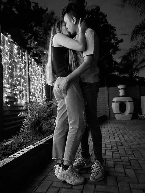 A Man And Woman Kissing In Front Of A Fence With Lights On The Wall