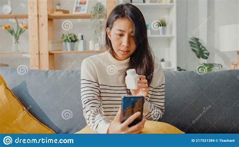 Sick Young Asian Woman Hold Medicine Sit On Couch Video Call With Phone Consult With Doctor At