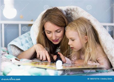 Young Mother Reading A Book Using Flashlight With Her Pretty Girl While Lying Down On Bed Stock