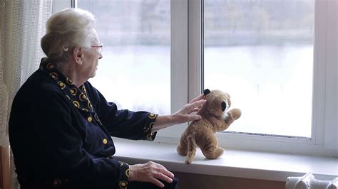 A Lonely Old Woman Looking Out The Window With A Soft Toy On The