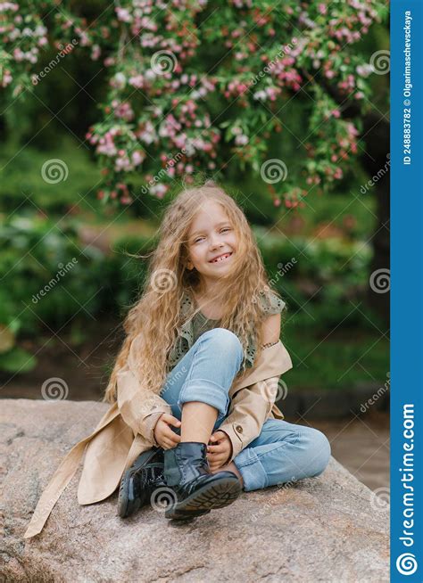uma doce e bela menina de sete anos e sentada numa rocha na primavera no jardim em uma capa de