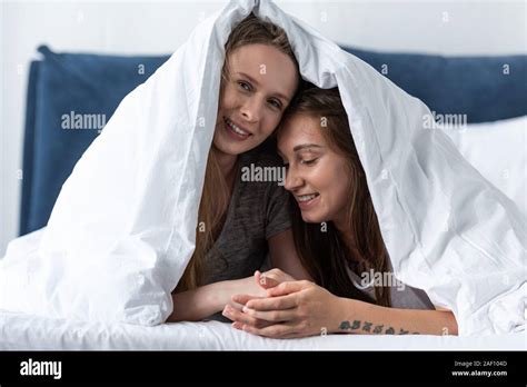 Two Happy Lesbians Holding Hands While Lying Under Blanket On Bed Stock