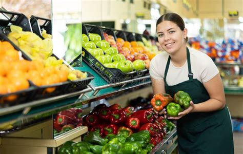 Positive Girl Seller In An Apron Offers Fresh Bell Peppers Stock Photo